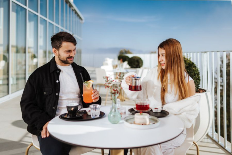 A man and a woman are sitting outside at a round table. They are drinking tea and juice. The woman is pouring tea, and the man is smiling. In the background, there is a view and a glass wall of the building.