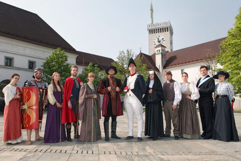 Moški in ženske v srednjeveških oblačilih stojijo pred Ljubljanskim gradom