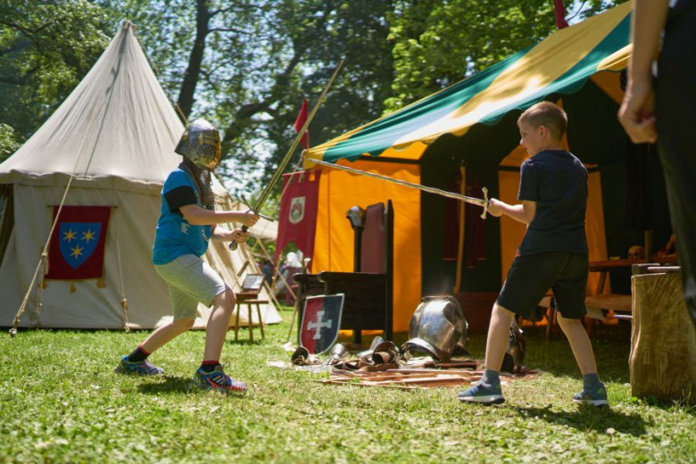 Two boys in medevial clothes in the front of the tents are fighting with sabers