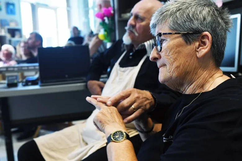 An older woman and a man in the foreground communicate through touch, using deafblind language. Other people can be seen in the background.