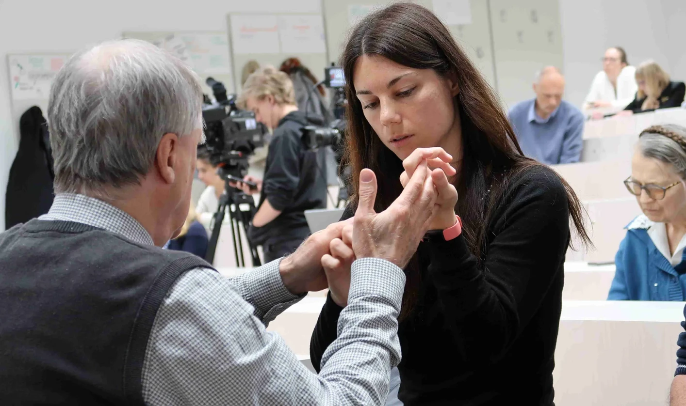 An older man and a young woman communicate through touch, using hand movements associated with deafblind language, while other people are present in the room.