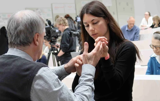 An older man and a young woman communicate through touch, using hand movements associated with deafblind language, while other people are present in the room.