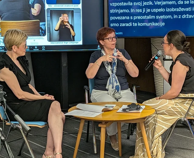 Three event participants take part in a discussion in a conference room. They are seated around a small table, with one participant speaking into a microphone and presentation screens visible in the background.