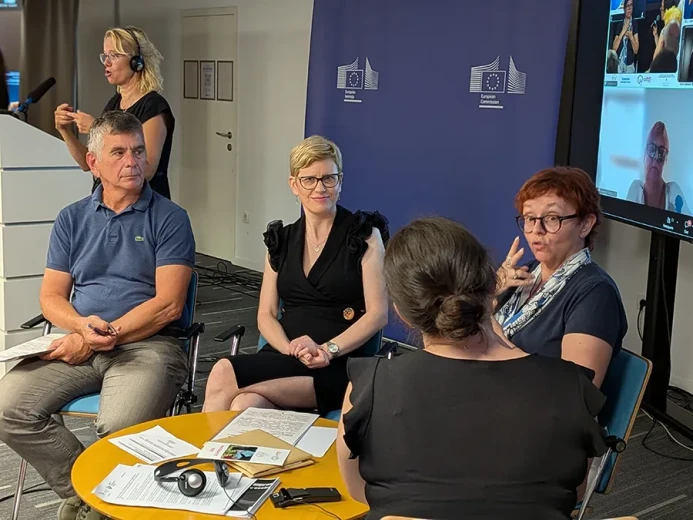 Participants in a panel discussion are seated around a round table at an event in a conference room. European Commission branding, presentation screens, and sign language interpretation are visible in the background.