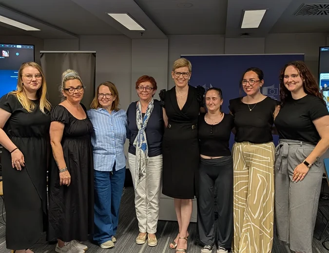 Group photo of eight event participants in a conference room. The participants are standing in a row in front of promotional display banners.