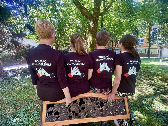 Four interpreters for deafblind people are seated on a bench in a park with their backs facing the camera. Their shirts display the text “Interpreter for Deafblind People” and a graphic illustrating hand-to-hand communication.