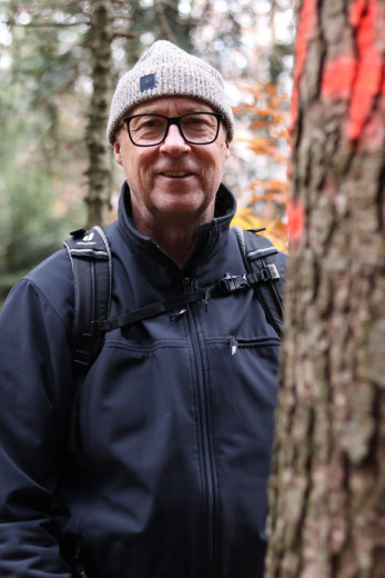 Tom Levanič wearing glasses, a beanie, and a jacket stands outdoors beside a tree with red markings.