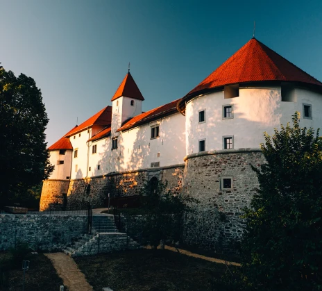 A historic white stone castle with red-tiled roofs and round towers stands against a clear blue sky, surrounded by trees.