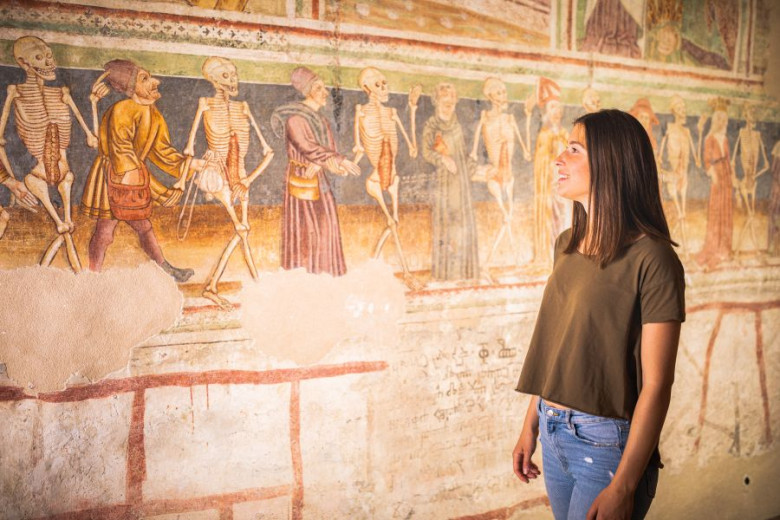 Ženska gleda stensko fresko s prikazom okostnjakov v zgodovinskem poslikanem prizoru.