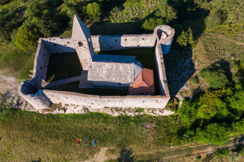 Pogled z zraka na kamniti grad s stolpi in obzidjem, obdan s travniki in drevesi.