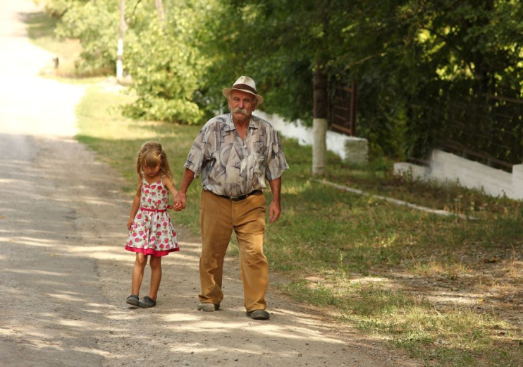 Dedek oziroma star človek in mala deklica se držita za roko in hodita med drevesi po neasfalitirani cesti