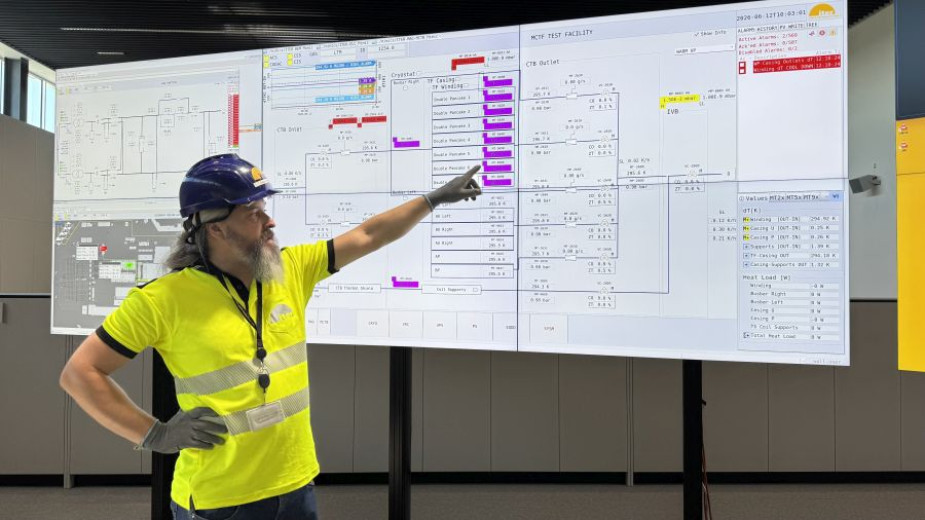 Gašper Renko, wearing a yellow shirt and a helmet, points at a large, complex control panel with multiple diagrams and data displays.