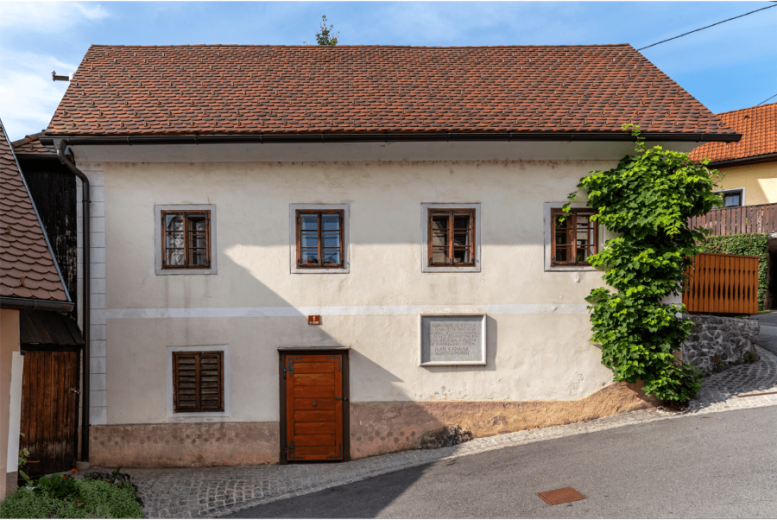 A quiet historic village house with a weathered white façade, red-tiled roof, wooden windows and door, and leafy vines beside a sloping road.