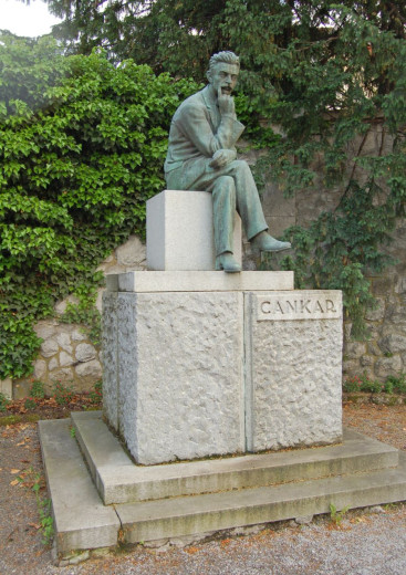 A contemplative bronze statue of a seated man rests on a pale stone pedestal engraved “CANKAR,” amid ivy and trees in a quiet garden.