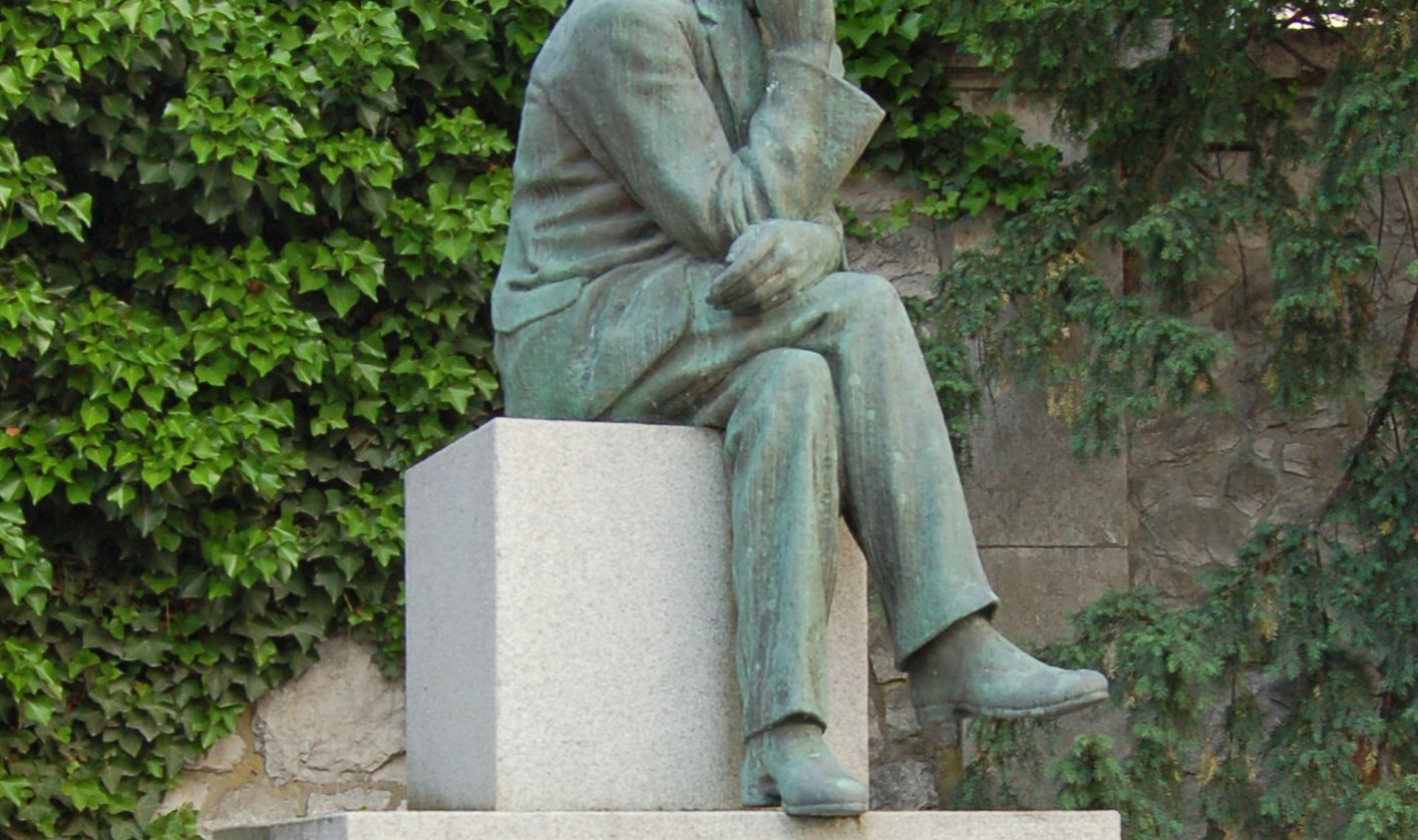 A contemplative bronze statue of a seated man rests on a pale stone pedestal engraved “CANKAR,” amid ivy and trees in a quiet garden.
