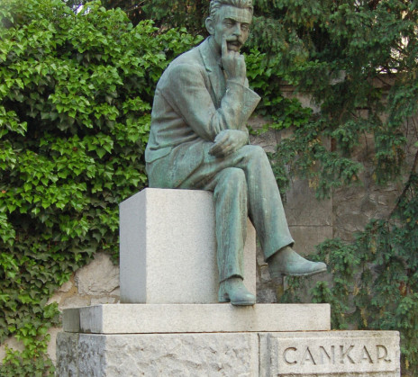 A contemplative bronze statue of a seated man rests on a pale stone pedestal engraved “CANKAR,” amid ivy and trees in a quiet garden.