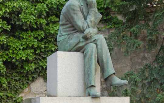 A contemplative bronze statue of a seated man rests on a pale stone pedestal engraved “CANKAR,” amid ivy and trees in a quiet garden.