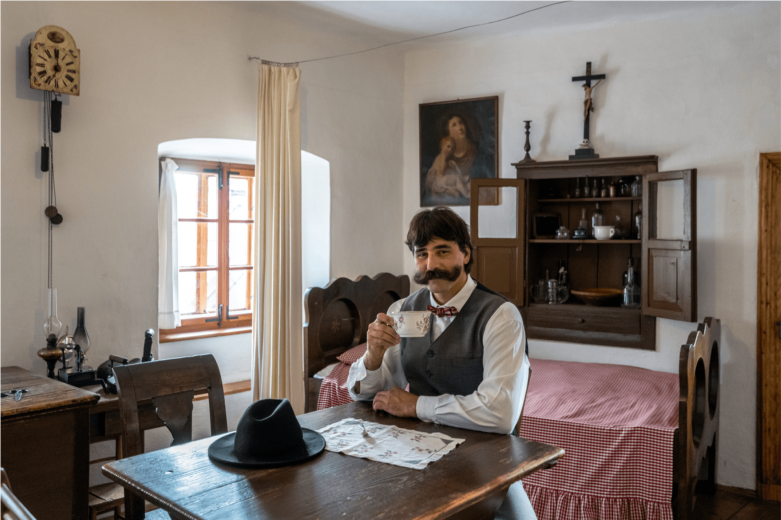 A mustached man in a vest sits at a wooden table, holding a small cup in a rustic room with a bed, cabinet, crucifix, and window; calm, nostalgic mood.