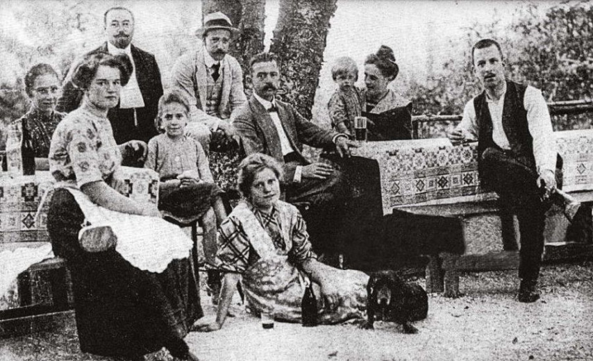 Grainy black-and-white archival photo of men, women, and a child gathered around a patterned outdoor table, with a dog at their feet; solemn mood.