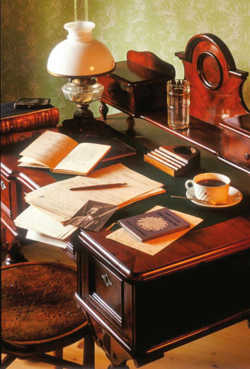 An antique wooden writing desk holds open books, handwritten papers, a pen, stacked volumes, and a teacup, lit by a warm oil lamp.