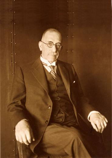 A sepia-toned portrait of Albert Bois de Chesne in a formal suit and round glasses, seated against a metallic backdrop.