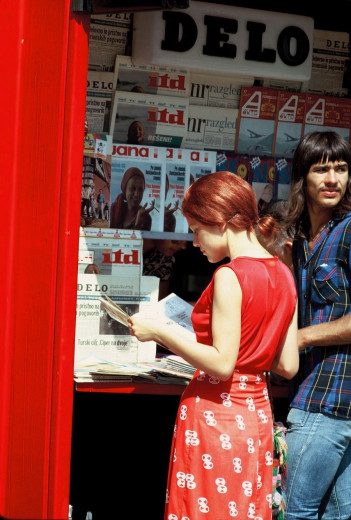 A woman in a red dress reads a newspaper at a box-style press kiosk covered with headlines, while a man behind her looks toward the street.