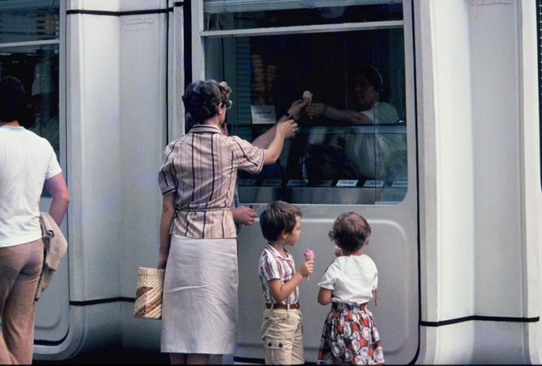 A woman with two small children stands by a train door, passing something through the window of a box-like service kiosk built into the carriage.