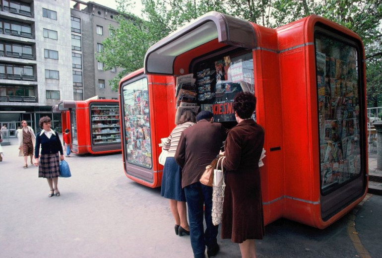 People queue at a bright red kiosk/newsstand packed with magazines; apartment blocks and trees line the street behind.