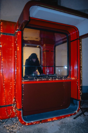 A bright red box-shaped kiosk glows with string lights at night, and a person inside works at a counter behind a large window.