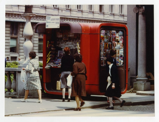 Several women walk and stop at a bright red box-shaped newspaper kiosk, whose open window is filled with magazines and daily headlines.