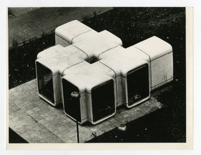 A black-and-white photo shows a cluster of connected box-shaped kiosk units forming a modular cube-like structure with large window panels.