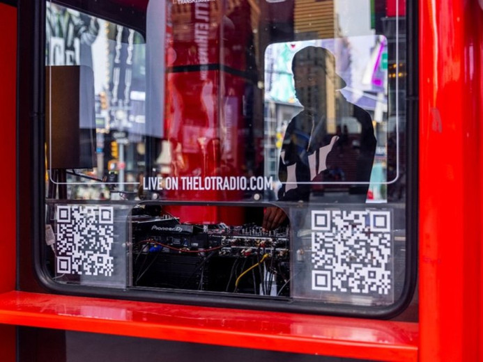 A red box-shaped kiosk has a glass window with two QR codes, and a person inside stands behind audio equipment and cables.
