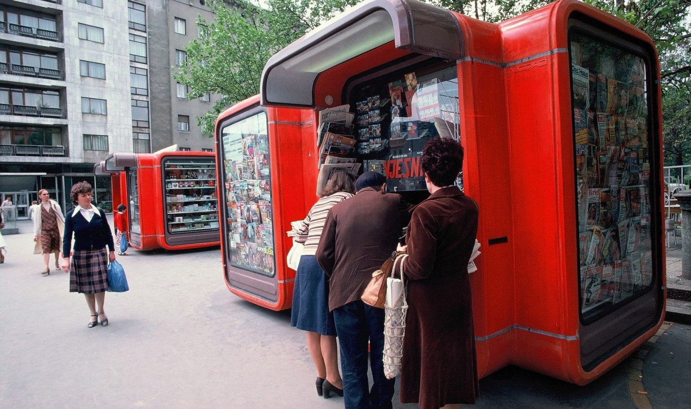 Three people stand at a bright red street kiosk filled with newspapers and magazines, with more kiosks and pedestrians along the city sidewalk.