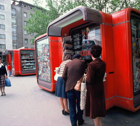 Three people stand at a bright red street kiosk filled with newspapers and magazines, with more kiosks and pedestrians along the city sidewalk.