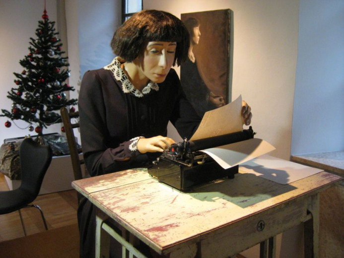 A wax figure of a woman, sitting behind a desk, typing on an old typewriter. 
