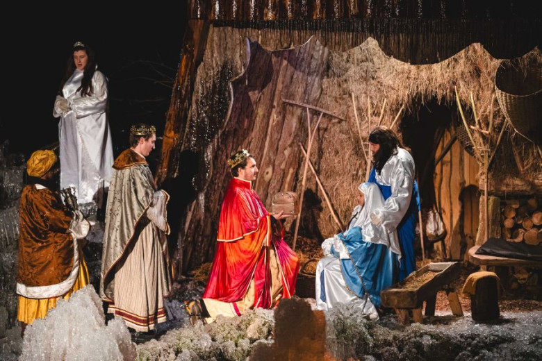 Live nativity scene with Mary and Joseph beside a manger in a rustic stable. The Three Wise Men in colorful robes kneel and stand before them, offering gifts. An angel in white watches from the side. The scene is set among straw, wood, and ice formations.