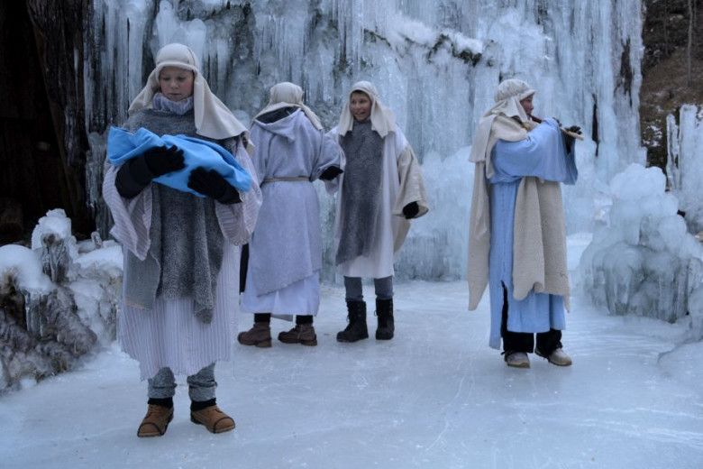 Shepherds in simple robes stand on icy ground before a wall of frozen cascades. One cradles a wrapped infant, while another plays a flute. Two others stand nearby, creating a quiet winter nativity scene.