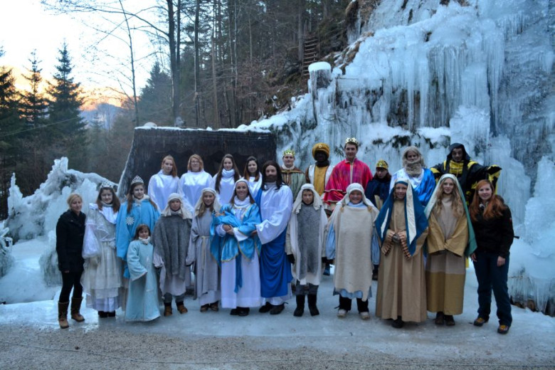 Group photo of performers in nativity costumes standing before icy cascades. Mary, Joseph, shepherds, angels, and the Three Wise Men pose together, with organizers beside them. The scene blends winter landscape and festive attire.