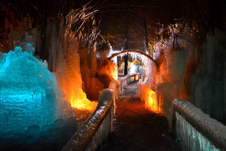 Ice tunnel illuminated with blue and warm orange light. Frozen walls and arches create a sculpted winter corridor leading into the distance, with reflections glowing on the icy surfaces.