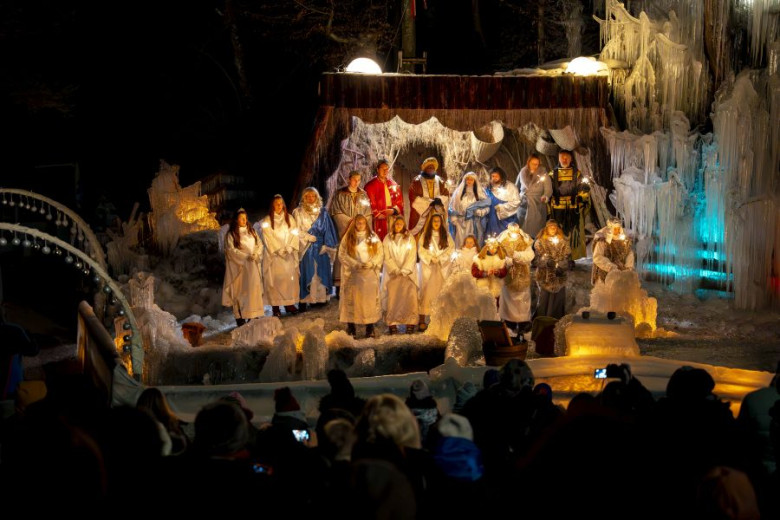 Outdoor performance of a live nativity scene on an icy stage. Actors dressed as Mary, Joseph, angels, shepherds, and the Three Wise Men stand in formation, illuminated by warm lights. Frozen structures frame the scene as the audience watches.