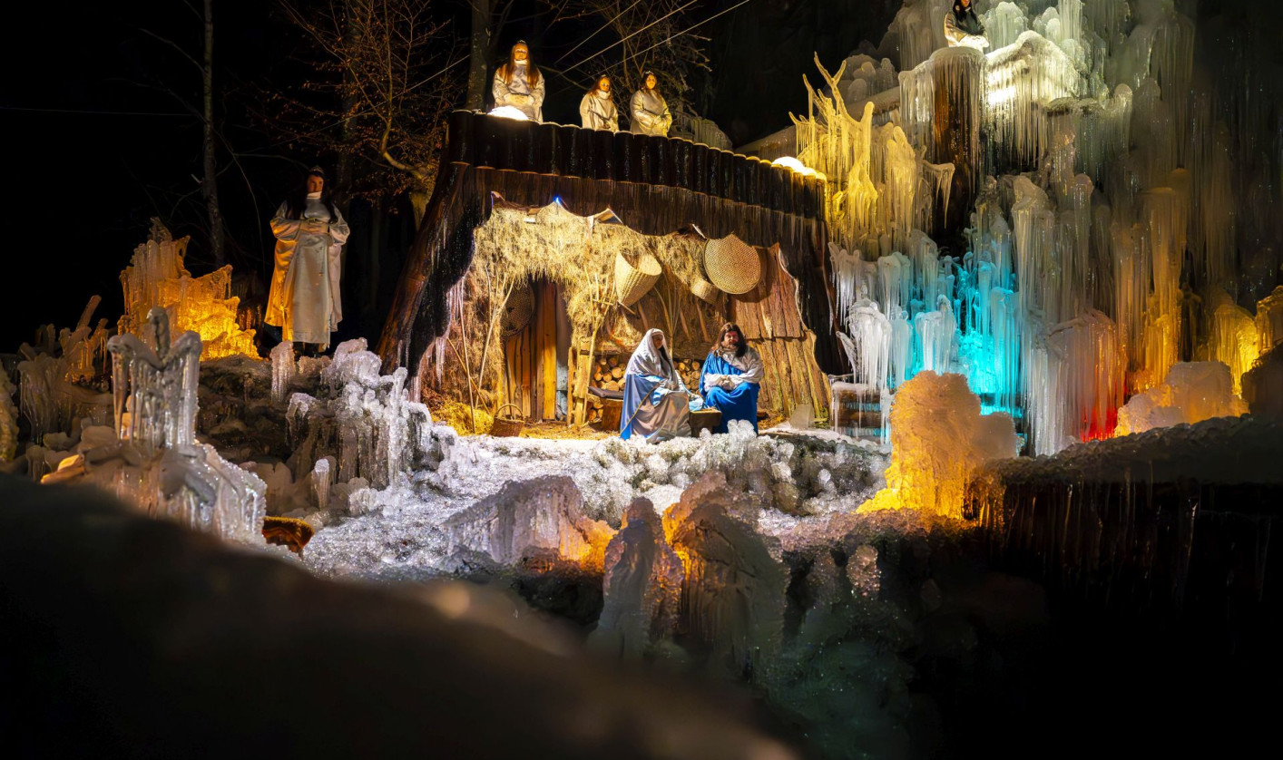 Outdoor performance of a live nativity scene on an icy stage. Actors dressed as Mary, Joseph, angels, shepherds, and the Three Wise Men stand in formation, illuminated by warm lights. Frozen structures frame the scene as the audience watches.