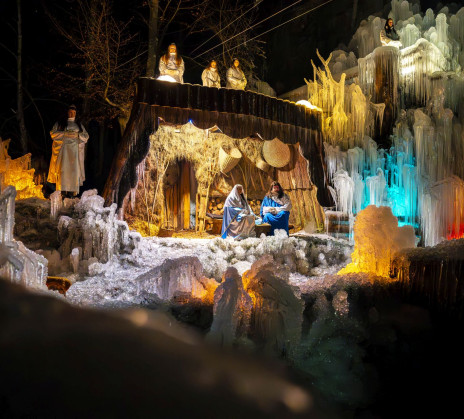 Outdoor performance of a live nativity scene on an icy stage. Actors dressed as Mary, Joseph, angels, shepherds, and the Three Wise Men stand in formation, illuminated by warm lights. Frozen structures frame the scene as the audience watches.