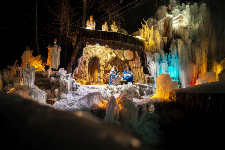 Outdoor performance of a live nativity scene on an icy stage. Actors dressed as Mary, Joseph, angels, shepherds, and the Three Wise Men stand in formation, illuminated by warm lights. Frozen structures frame the scene as the audience watches.
