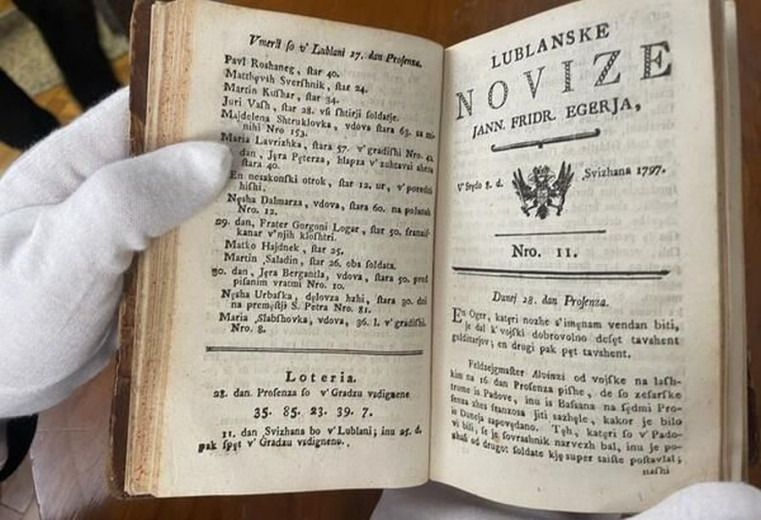 Old book "Lublanske Novize" from 1797, open, showing text and lottery numbers, held with white gloves.