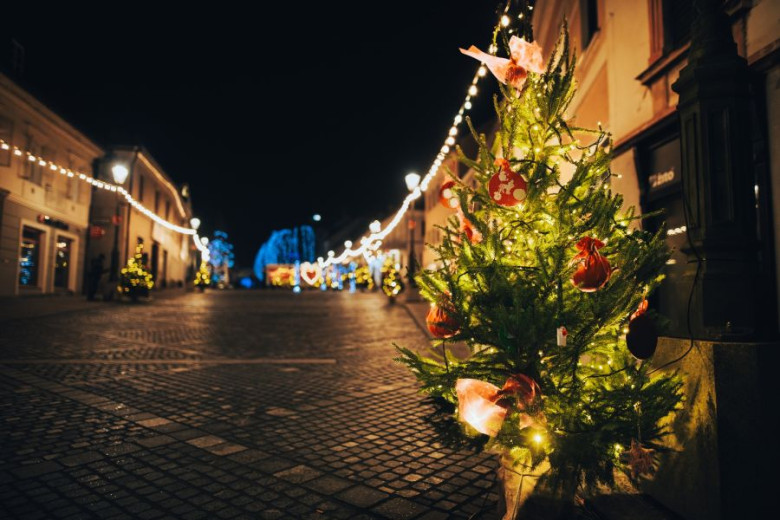 A street decorated with yellow lights, with a small Christmas tree in the foreground adorned with yellow lights and red ornaments.