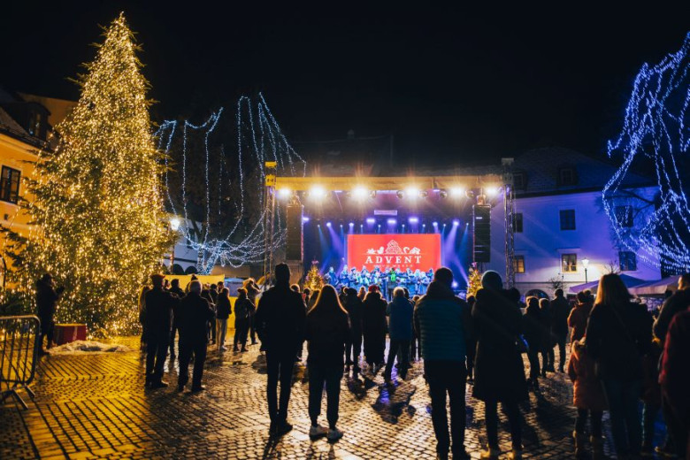 People stand in the square as a concert takes place on the stage, with a large, festively decorated Christmas tree on the right.
