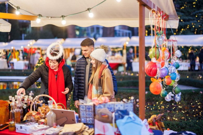 A young couple browses a holiday stall, where the vendor shows them items such as spirits and ornaments.