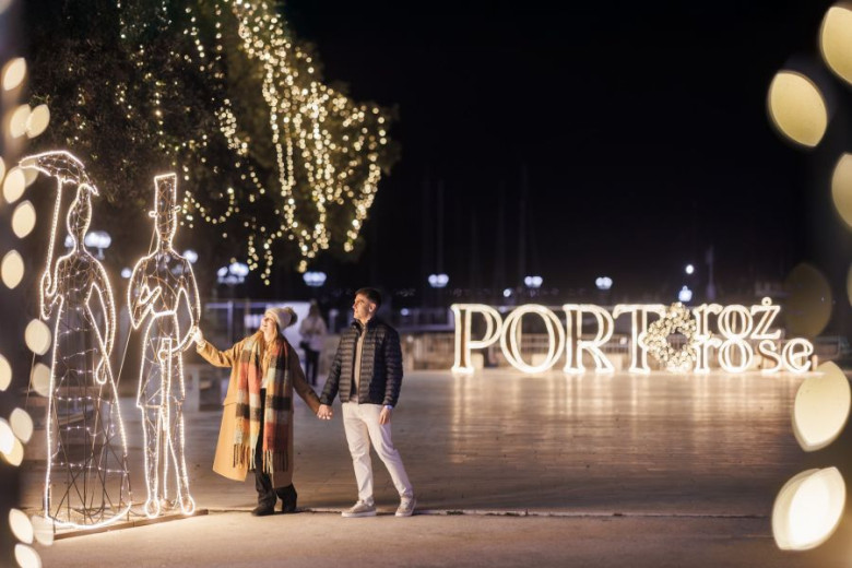 A young couple stands beside two figures made of holiday lights, with a glowing “Portorož” sign visible behind them.