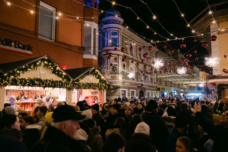 A crowd gathers on a festively decorated street with yellow lights and red baubles, with two holiday stalls on the left side offering seasonal treats and gifts.