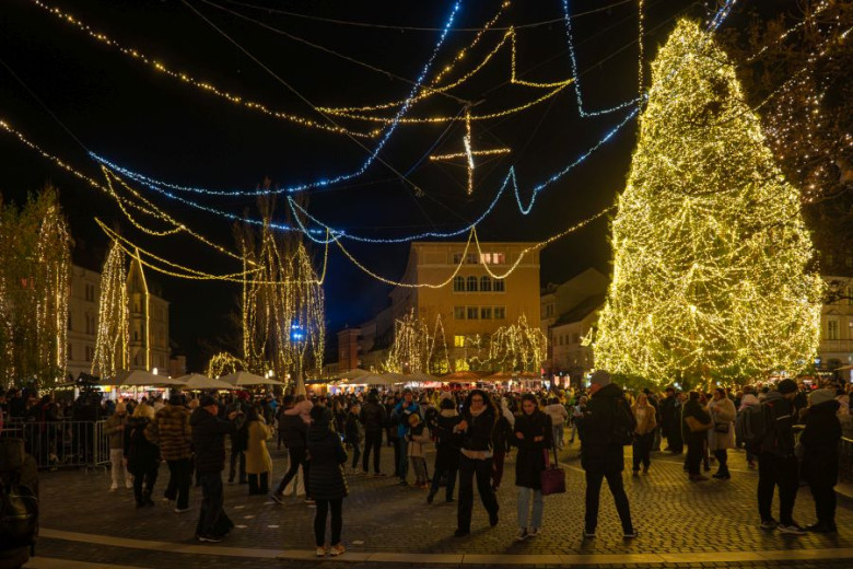 A crowd gathers at Prešeren Square in Ljubljana, decorated with festive lights, where a large Christmas tree stands adorned with thousands of tiny white lights.